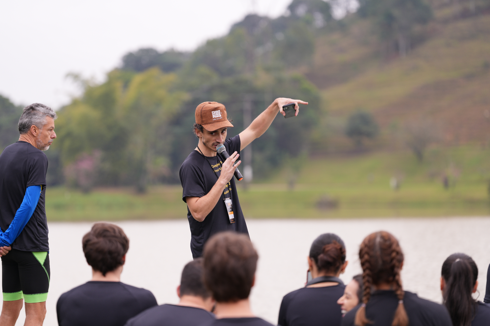 Ariel Perazza ao microfone apontando para o horizonte à beira do lago enquanto orienta os participantes da Airport Experience