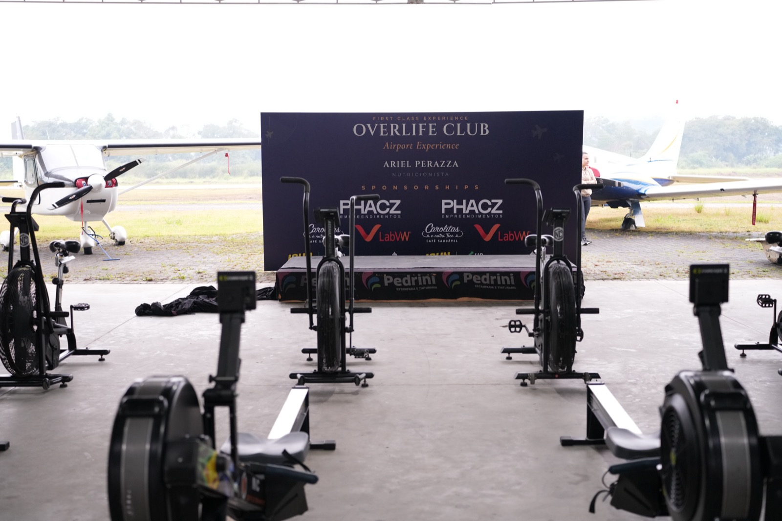 Estação Hybrid Hangar montada no hangar com remos e airbikes e o painel Overlife Club ao fundo