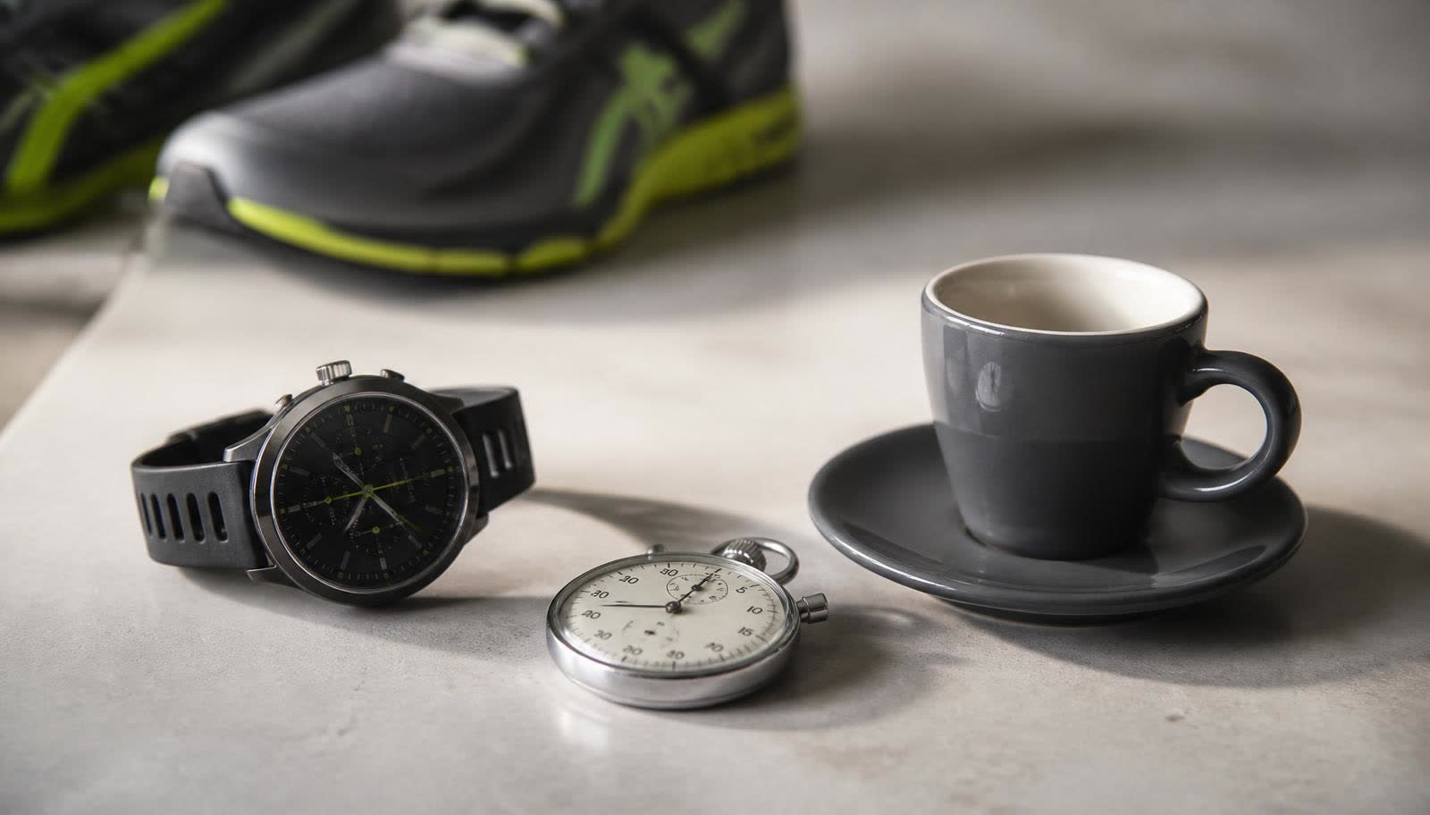 Relógio esportivo de pulso e cronômetro ao lado de uma xícara de café vazia sobre bancada clara, com tênis de corrida desfocado ao fundo, representando o timing da cafeína antes do treino