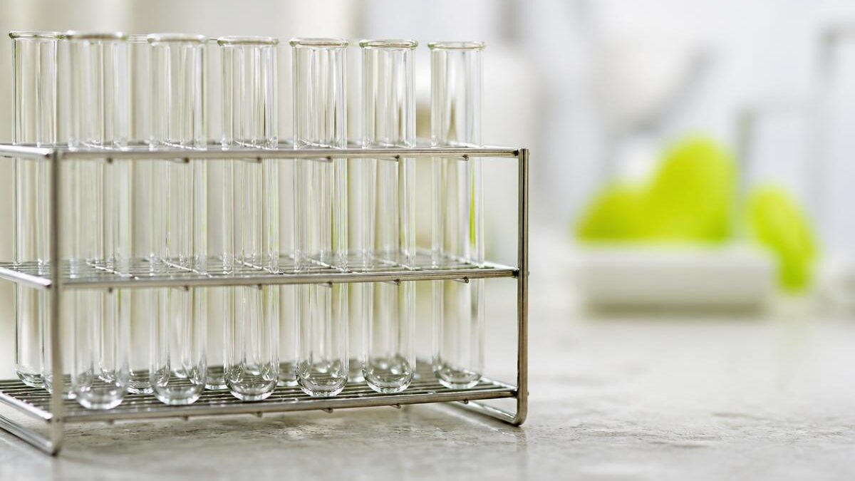 Close de tubos de ensaio de vidro transparentes e vazios em rack metálico sobre superfície de pedra clara, com profundidade de campo rasa e luz natural difusa