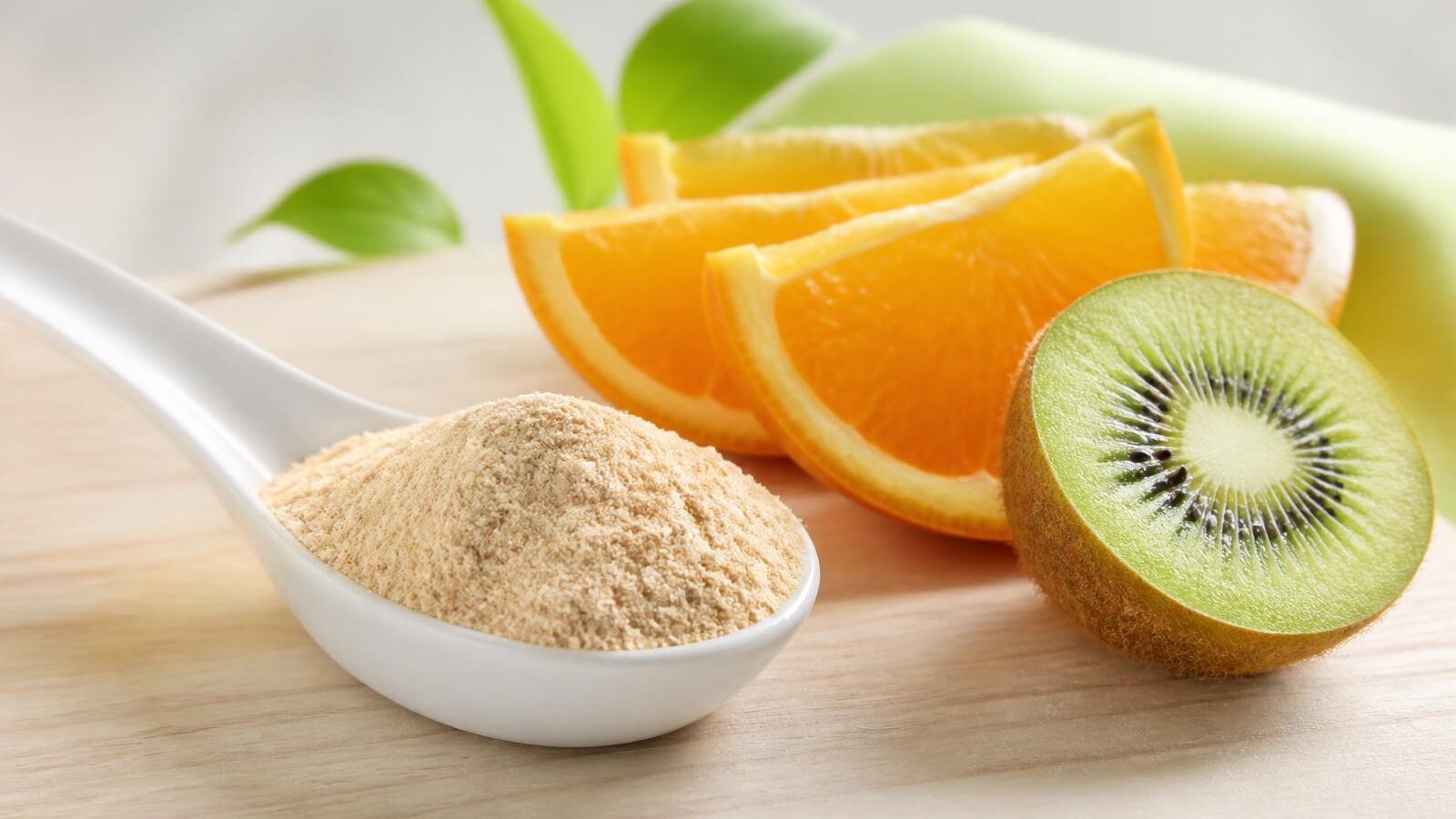 Colher branca cheia de pó bege claro sobre superfície de madeira clara ao lado de rodelas de laranja e kiwi cortado