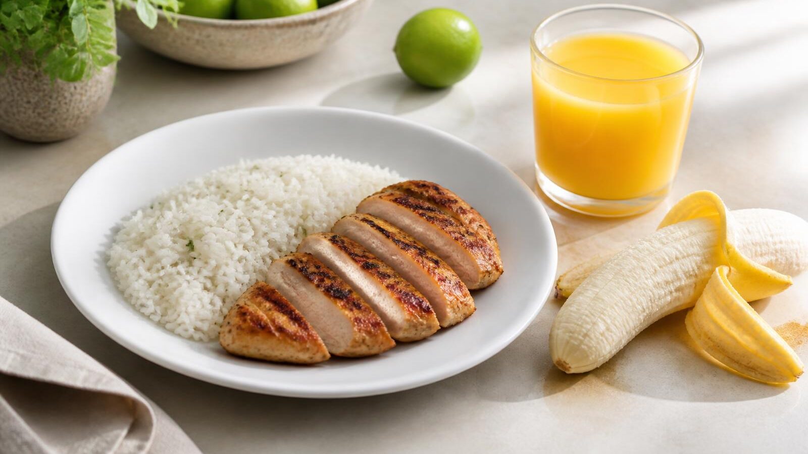 Prato branco com arroz branco e peito de frango grelhado fatiado sobre bancada clara, ao lado de uma banana descascada pela metade e um copo de suco de laranja