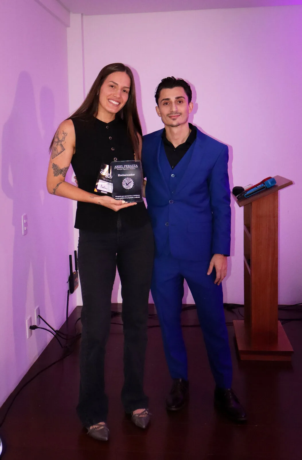 Embaixadora recebendo placa de reconhecimento ao lado de Ariel Perazza no Overlife Day 2025