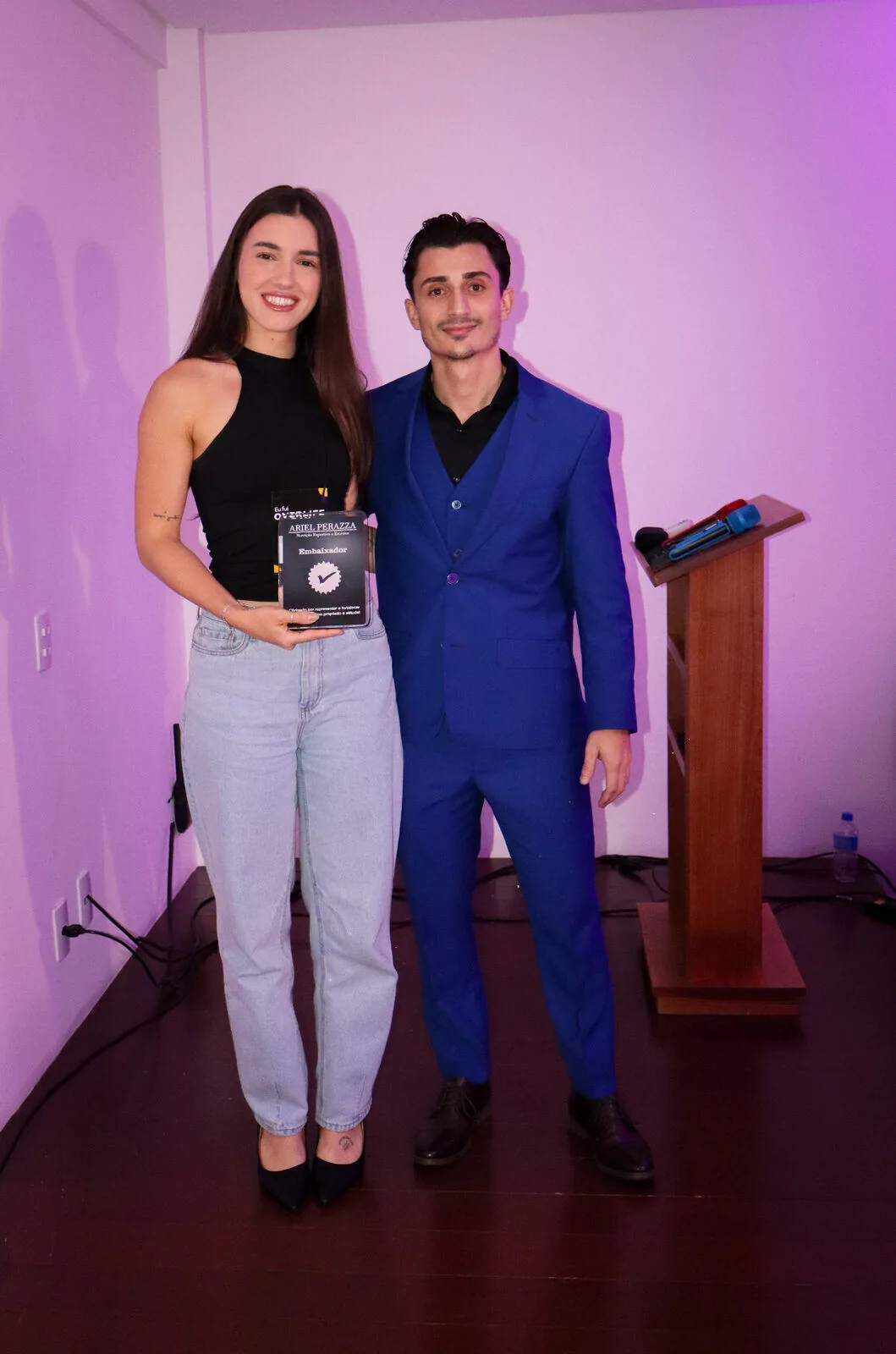 Embaixadora recebendo placa de reconhecimento ao lado de Ariel Perazza no Overlife Day 2025